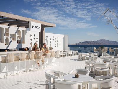 Ristorante all'aperto bianco moderno sul mare con tavoli, sedie e bar sotto un cielo azzurro.