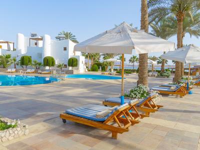 Sonnenschirme und Liegestühle neben einem Pool in einem Hotel mit weißen Gebäuden und Palmen.