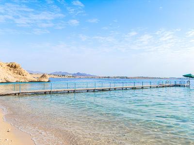 Langer Steg führt über klares türkisfarbenes Wasser an einem sonnigen Strand mit hellem Sand.