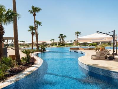 Ein tropischer Hotelpool mit Palmen, Liegestühlen und Sonnenschirmen bei klarem Himmel.