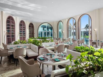 Helles Hotelrestaurant mit großen Rundbogenfenstern, bequemen Stühlen und grünen Pflanzen.