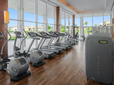 Heller Fitnessraum mit Holzfußboden, mehreren Laufbändern und großen Fenstern mit Blick auf Palmen.