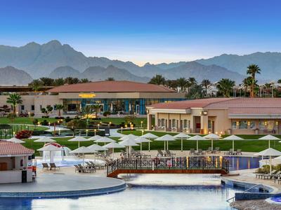 Luxuriöses Hotel mit Pool und Liegestühlen vor einer Bergkulisse bei klarem Himmel.