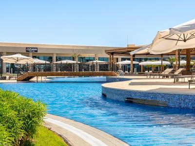 Großer Hotelpool mit blauer Umrandung, Liegestühlen und Sonnenschirmen unter klarem Himmel.