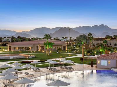 Hotel mit großem Pool, weißen Sonnenschirmen, Palmengarten und Bergen im Hintergrund bei Abenddämmerung.
