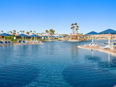 Großer Hotelpool mit Sonnenliegen, Sonnenschirmen und Palmen unter klarem blauem Himmel.
