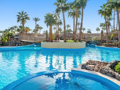 Großer Hotelpool mit Palmen und Sonnenterrassen unter klarem Himmel.