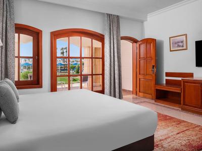 Helles Hotelzimmer mit großem Bett, hölzernen Fenstern und Tür, TV und Teppichboden.