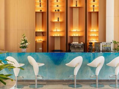 Area bar moderna in hotel con sgabelli bianchi e bancone in marmo verde-blu sotto mensole illuminate.