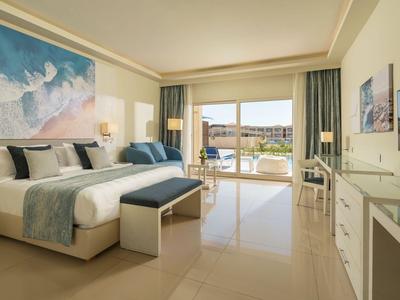 Camera d'albergo luminosa con letto grande, panca ai piedi e vista su balcone e piscina.