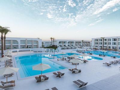 Moderner Hotelpoolbereich mit Liegestühlen, Sonnenschirmen und weißen Gebäuden unter blauem Himmel.