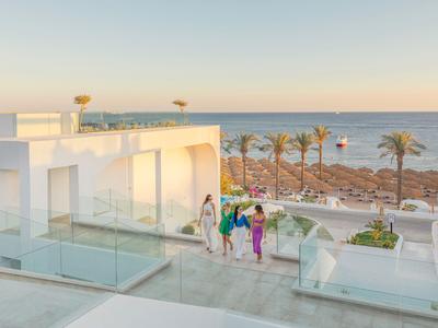 Moderne weiße Hotelterrasse mit Glasgeländer, drei Personen und Blick aufs Meer bei Sonnenuntergang.