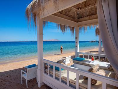 Blick auf einen Sandstrand mit klarblauem Wasser und einer Lounge-Hütte mit weißen Möbeln.