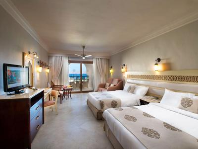 Helles Hotelzimmer mit zwei Betten, TV, Schreibbereich und Blick auf Meer durch Fenster.