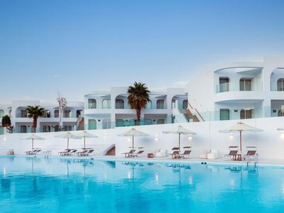 Hotel bianco con palme e ombrelloni accanto alla grande piscina blu sotto un cielo limpido.