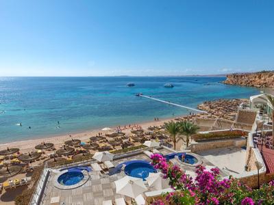 Blick auf einen Strand mit Liegestühlen, Pools und blühenden Blumen unter klarem Himmel.