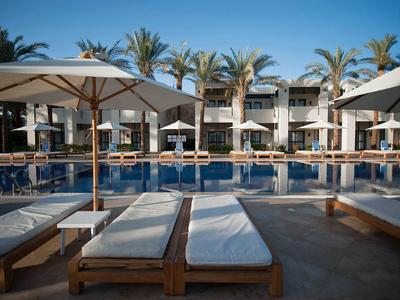 Modern pool area with loungers and umbrellas in front of a hotel with palm trees in the background.
