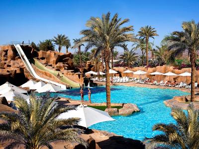 Großer Pool mit Wasserrutschen, Palmen und Sonnenschirmen in einer Urlaubsanlage.