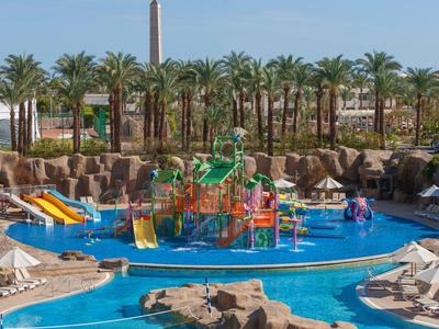 Wasserpark mit buntem Klettergerüst und Wasserrutsche, umgeben von Palmen und Liegestühlen.
