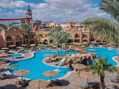 Piscina de hotel con juegos de agua azules, sombrillas y palmeras bajo un cielo nublado.