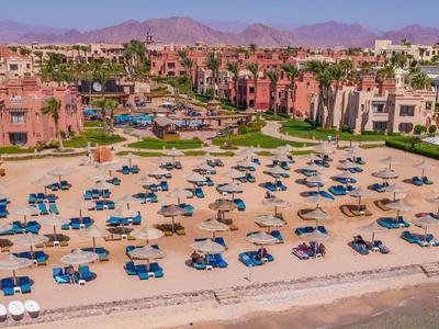 Ein breiter Sandstrand mit blauen Liegestühlen vor einem Resort mit mehreren Gebäuden und Pools.