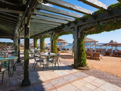 Terraza cubierta con sillas y mesas de metal con vistas a una playa de arena y al mar bajo el sol.