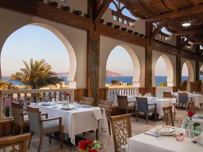 Terraza de restaurante con mesas blancas y vista al mar a través de grandes arcos a la luz del día.