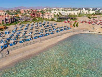 Blick auf einen Strand mit blauen Sonnenschirmen und klarem Meer vor Ferienanlagen.