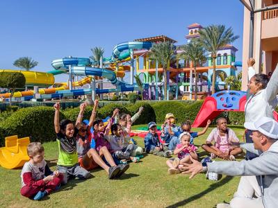 Kinder sitzen lachend auf Gras vor einem farbenfrohen Wasserpark mit Rutschen und Palmen.