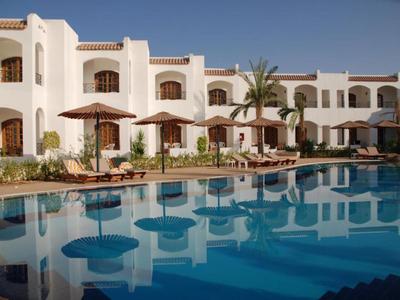 Hotel mit weißer Fassade und großem Pool, umgeben von Sonnenschirmen und Palmen.