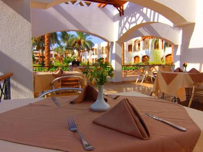 Ein gedeckter Tisch mit Besteck und dreieckiger Serviette vor einem sonnigen Hotelrestaurant mit tropischer Umgebung.