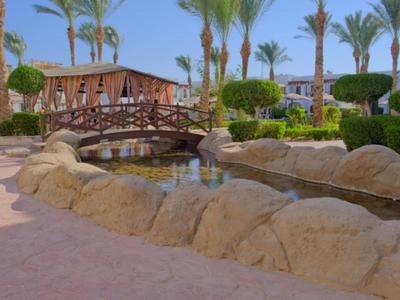 Ein Garten mit Palmen, einem Steg über einem Wasserlauf und Holzgebäuden im Hintergrund.
