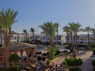 Hotelanlage mit Pool, Palmen, Pavillons und weißer Architektur bei klarem Himmel.