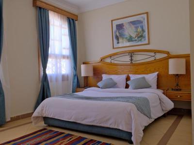 Helles Hotelzimmer mit großem Bett, Holzkopfteil, blauen Vorhängen und buntem Teppich.