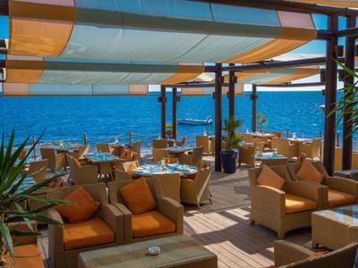 Offene Terrasse mit bequemen Sesseln und Tischen mit Blick auf das blaue Meer unter Sonnensegeln.