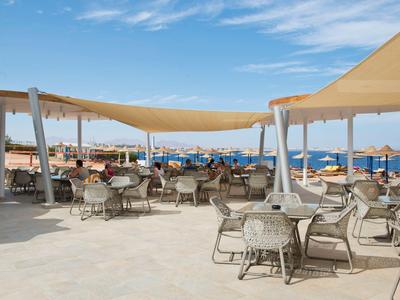 Offener Sitzbereich mit Tischen und Stühlen unter Sonnensegeln am Strand bei klarem Wetter.