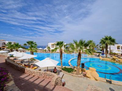 Hotelpool mit Palmen, Sonnenschirmen und weißen Gebäuden unter leicht bewölktem Himmel.