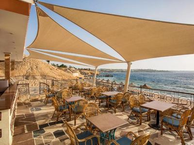 Terrasse mit Tischen und Stühlen unter Sonnensegeln mit Blick auf das Meer bei Sonnenuntergang.