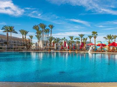 Großer Hotelpool bei Sonnenschein mit Palmen, roten Sonnenschirmen und moderner Architektur.