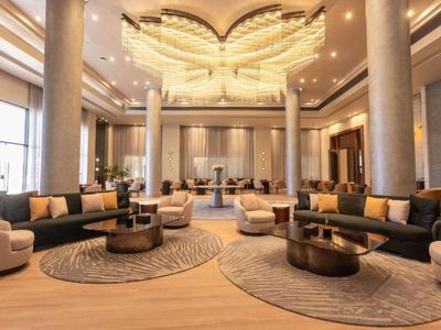 Przestronny hol hotelowy z wysokimi kolumnami, okrągłymi dywanami i nowoczesnymi miejscami do siedzenia w ciepłych odcieniach.