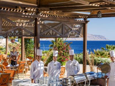 Freiluftrestaurant mit Kellnern, Gästen und Blick auf das blaue Meer und den Himmel.