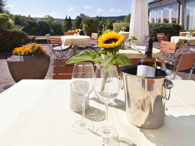 Tisch mit Weingläsern, Sektkühler und Sonnenblume auf Terrasse mit Stühlen und Pflanzen im Hintergrund.