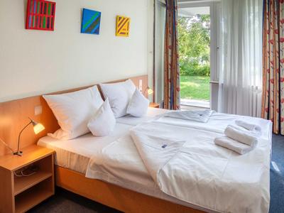 Helles Hotelzimmer mit Doppelbett, Nachttisch, Wandkunst und Blick in den Garten.