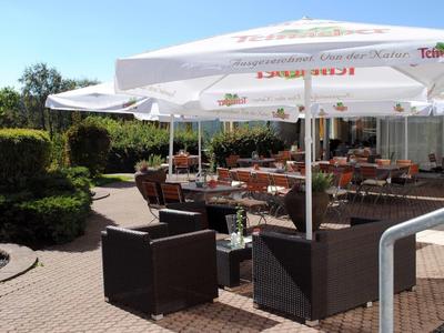 Außenbereich eines Cafés mit weißen Sonnenschirmen, braunen Sitzmöbeln und Pflanzen im Hintergrund.