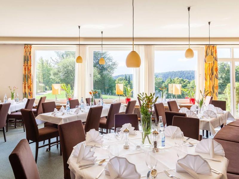 Hell eingerichteter Speisesaal mit rund gedeckten Tischen, braunen Stühlen und großen Fenstern.