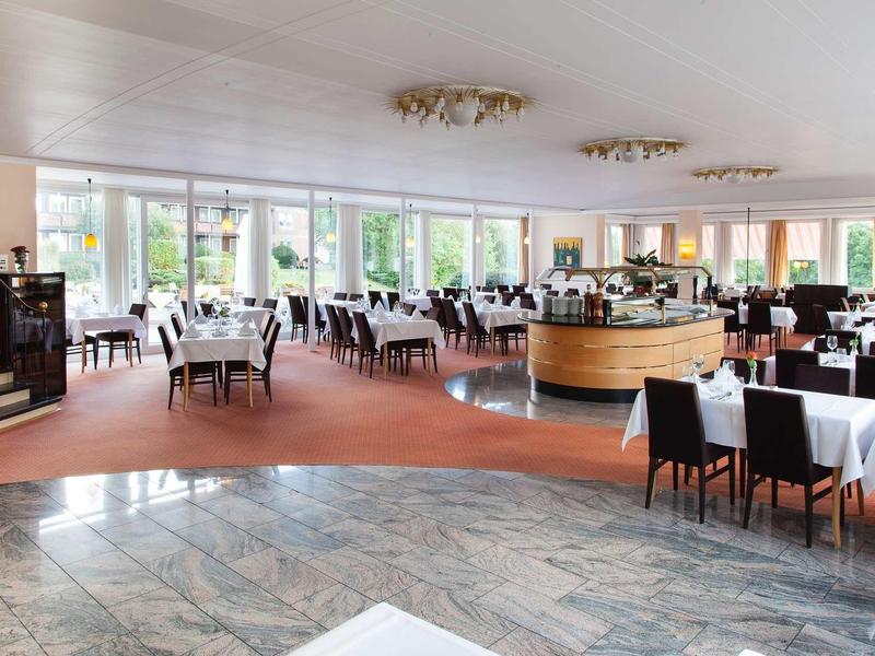 Großer, heller Restaurantraum mit weißen Tischdecken, schwarzen Stühlen und bodentiefen Fenstern.