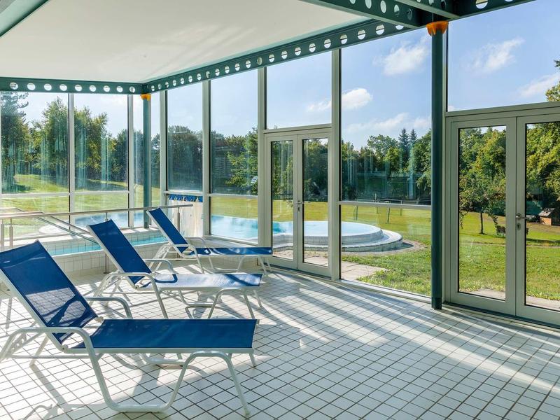 Geschlossener Poolbereich mit blauen Liegestühlen, großen Glasfenstern und Blick auf grünen Garten.