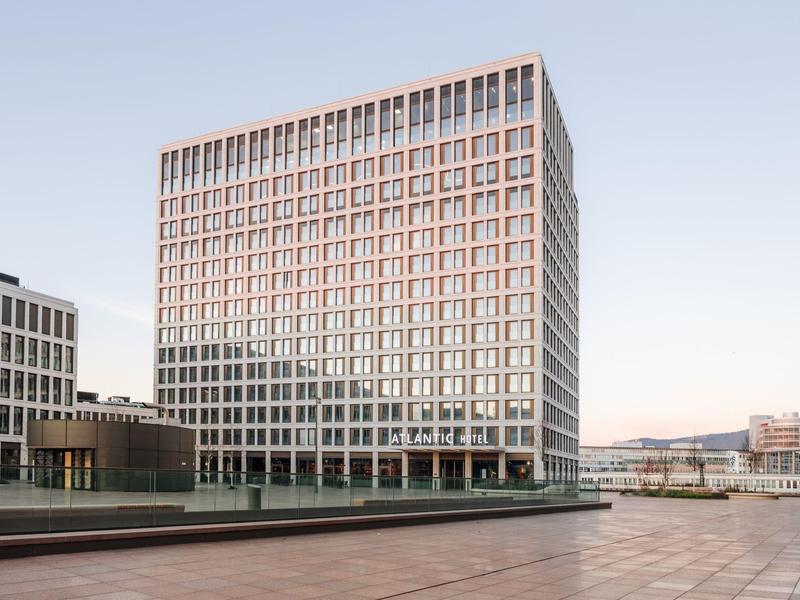 Modernes Bürogebäude mit Glasfenstern und quadratischer Form in urbaner Umgebung bei klarem Himmel.