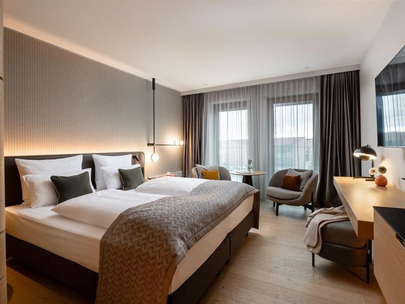 Modernes Hotelzimmer mit großem Bett, zwei Sesseln, Wandfernseher und Fenster mit grauen Vorhängen.