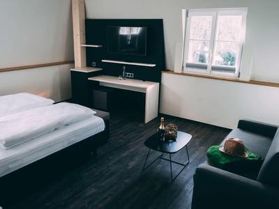 Kleines Zimmer mit Bett, Fernseher, schwarzem Sofa und rundem Tisch, helle Wände und Fenster.